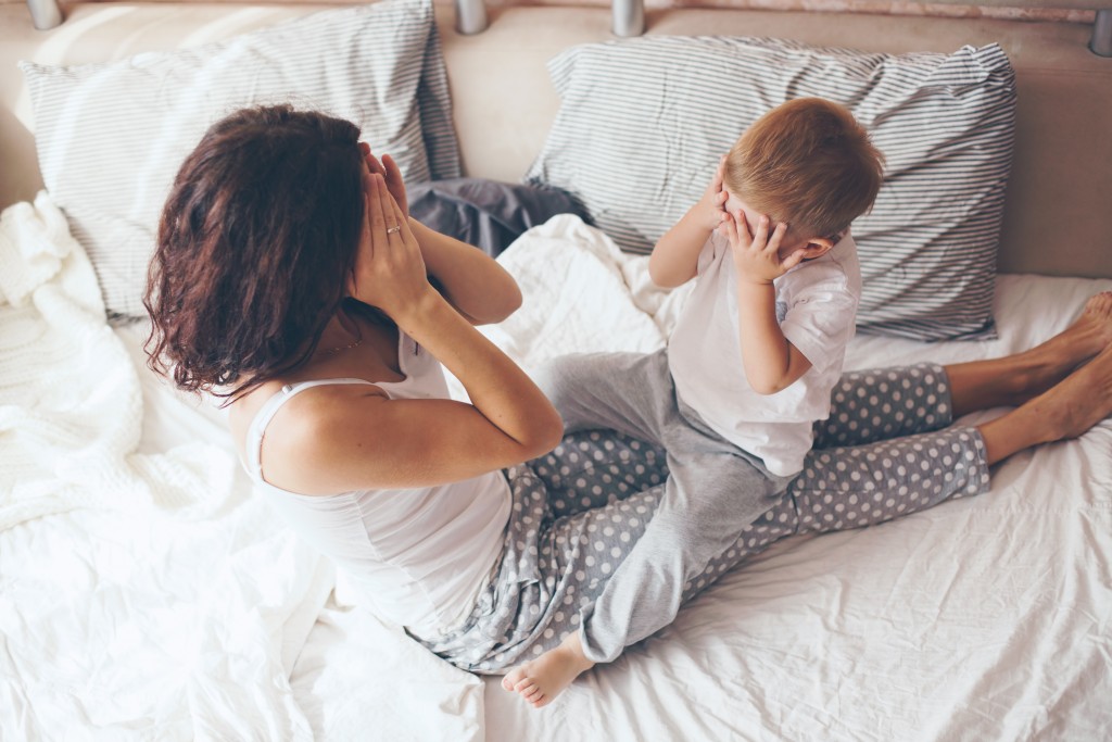 Young mother with her 2 years old little son dressed in pajamas are relaxing and playing in the bed at the weekend together, lazy morning, warm and cozy scene. Pastel colors, selective focus.